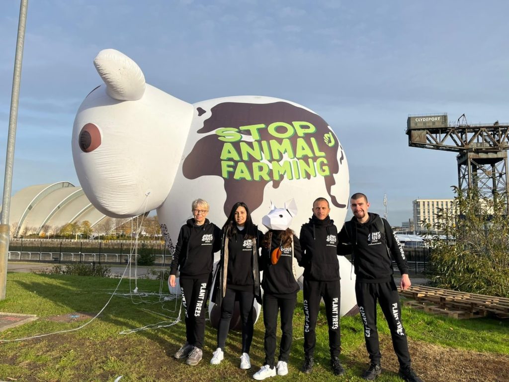 Five animal rights activists wear black clothes with white slogans that read "plant trees" as they stand in front of a 10-ft tall inflatable cow with the message "stop animal farming" on the side. 