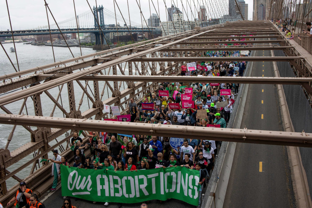 Hundreds march across a bridge in New York. They are protesting for women's reproductive rights and carrying a large green banner that reads "Our Abortions".