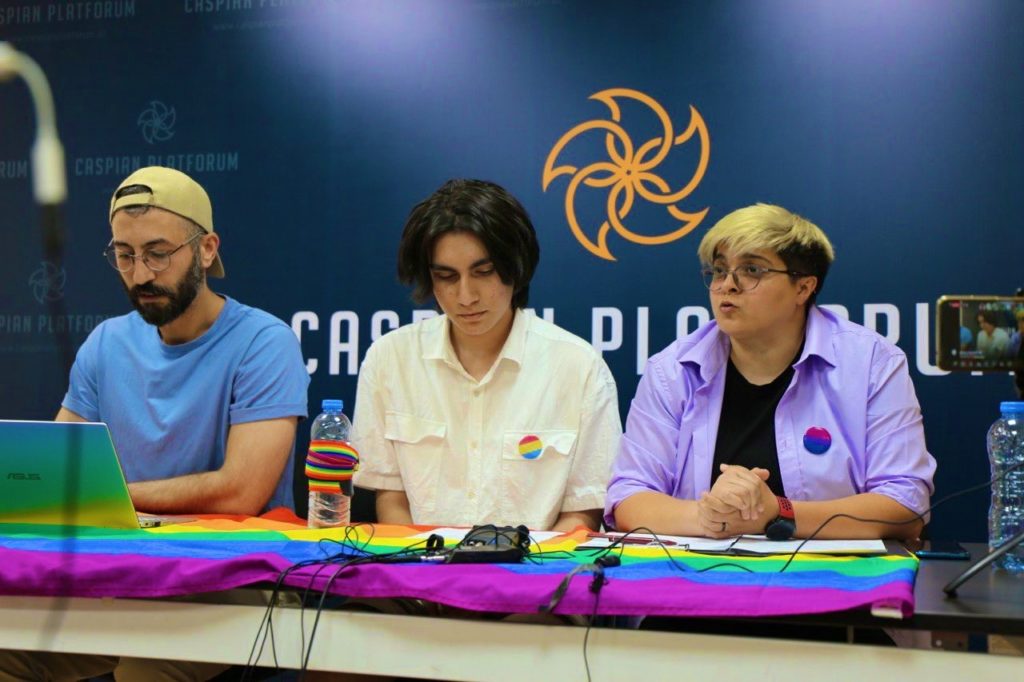 Three queer organizers hold a press briefing in Azerbaijan. A rainbow flag symbolizing LGBTQ+ rights is on the table. Protesting is risking for queer people in Azerbaijan.