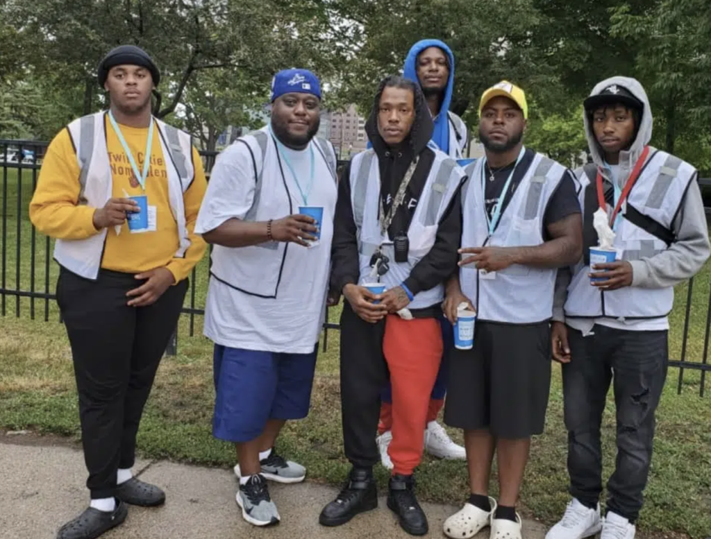 Wearing gray vests, five African-American men and their mentor stand near a neighborhood park in Minneapolis. They are community peacekeepers who stop violence with nonviolent de-escalation.