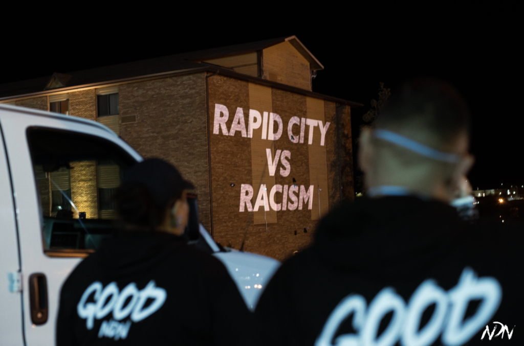 At night, two Native Americans project a light display protest on the front of a brick hotel in Rapid City, South Dakota. It reads, "Rapid City vs. Racism" and is part of a campaign to boycott a set of racist hotels. 