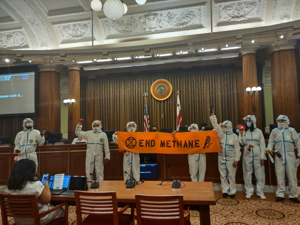 Wearing white hazmat suits and carrying an orange banner that reads "end methane", climate activists disrupt a city council meeting.