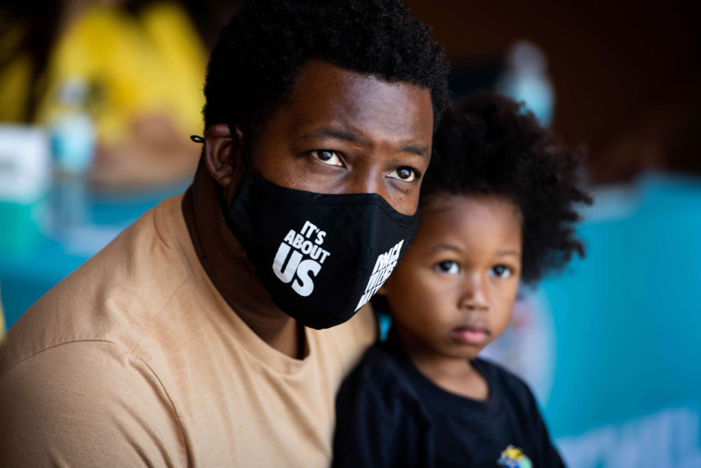 A Black father holds his son while wearing a mask that reads "it's about US". 