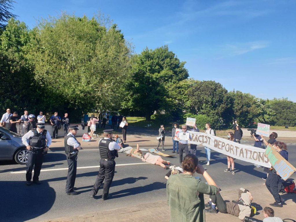 Protesters stretch banner across street and lay down on pavement to stop deportations of Rwandan asylum seekers in United Kingdom.