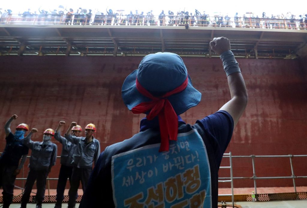 South Korean shipyard workers in hard hats and blue vests rally and chant inside the half-built ship they have occupied inside their factory.