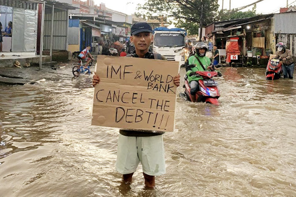 An Indonesian man stands in a flooded street holding a sign that says, "IMF & World Bank - Cancel the Debt".