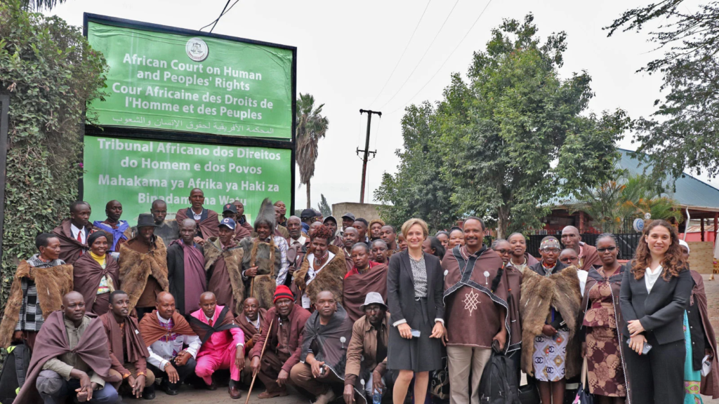 A large group of Indigenous Ogiek people stand in traditional garb in front of two large green billboards for the African Court on Human and Peoples' Rights.