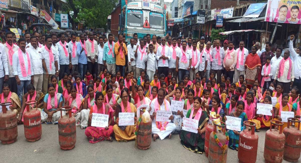 A crowd of Indian men stand behind a seated group of mostly women amidst a road block consisted of empty fuel canisters. They all wear pink scarves and are protesting high cooking fuel prices.