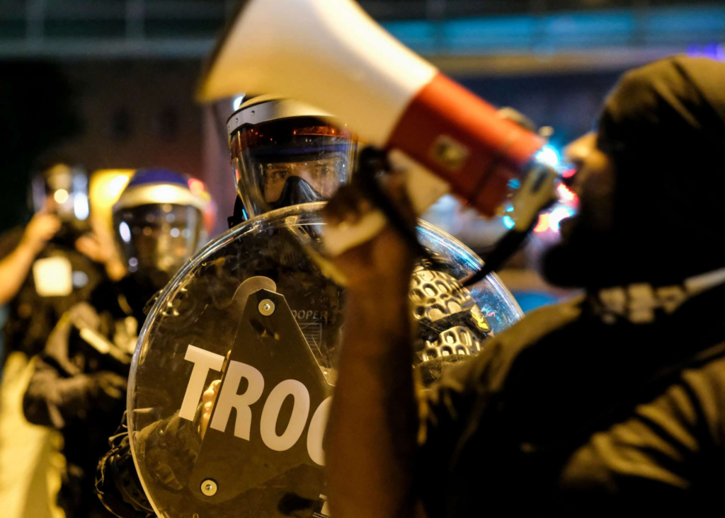A riot police officer holding a round shield stares at a Black man with a megaphone at a protest against the killing of Jayland Walker in Akron, OH.