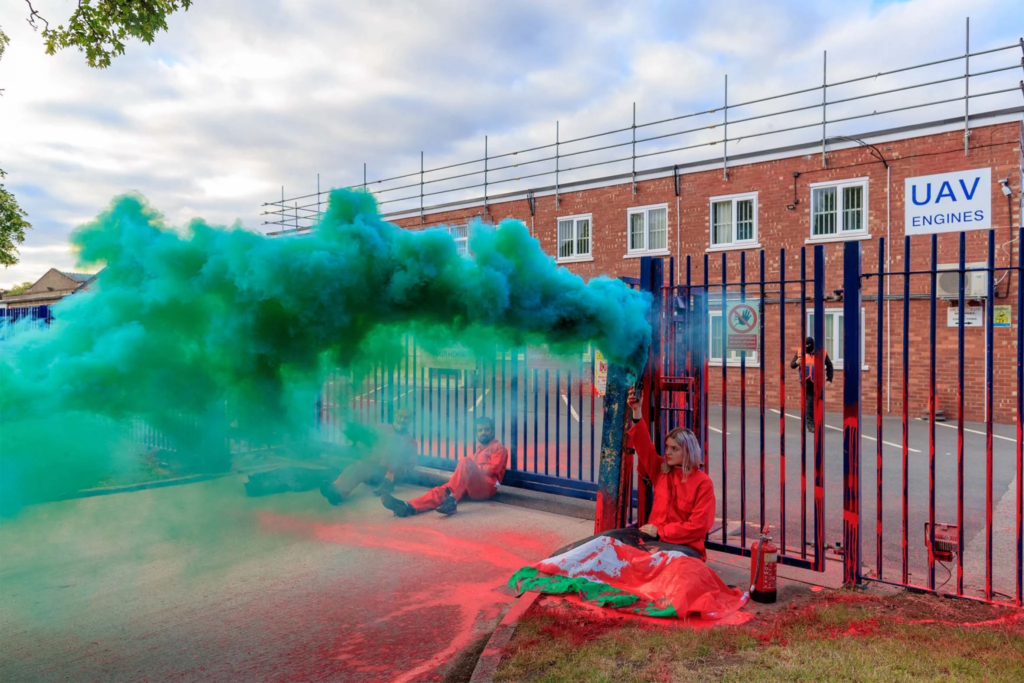 Three peace activists in bright red jumpsuits lock themselves to the gates of a weapons factory in the United Kingdom that manufactures weapons that Israel uses against Palestine. One activist holds aloft a teal smoke flare.