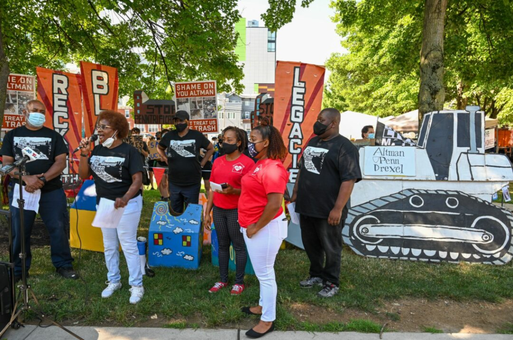 Six Black people stand together as a woman speaks into a microphone at a rally against luxury home development in Philadelphia.
