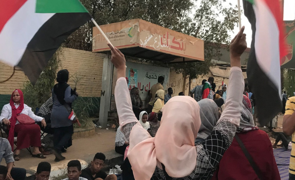 A Sudanese woman wearing a pink headscarf holds two national flags aloft at a street demonstration during the nonviolent resistance to the dictatorship.