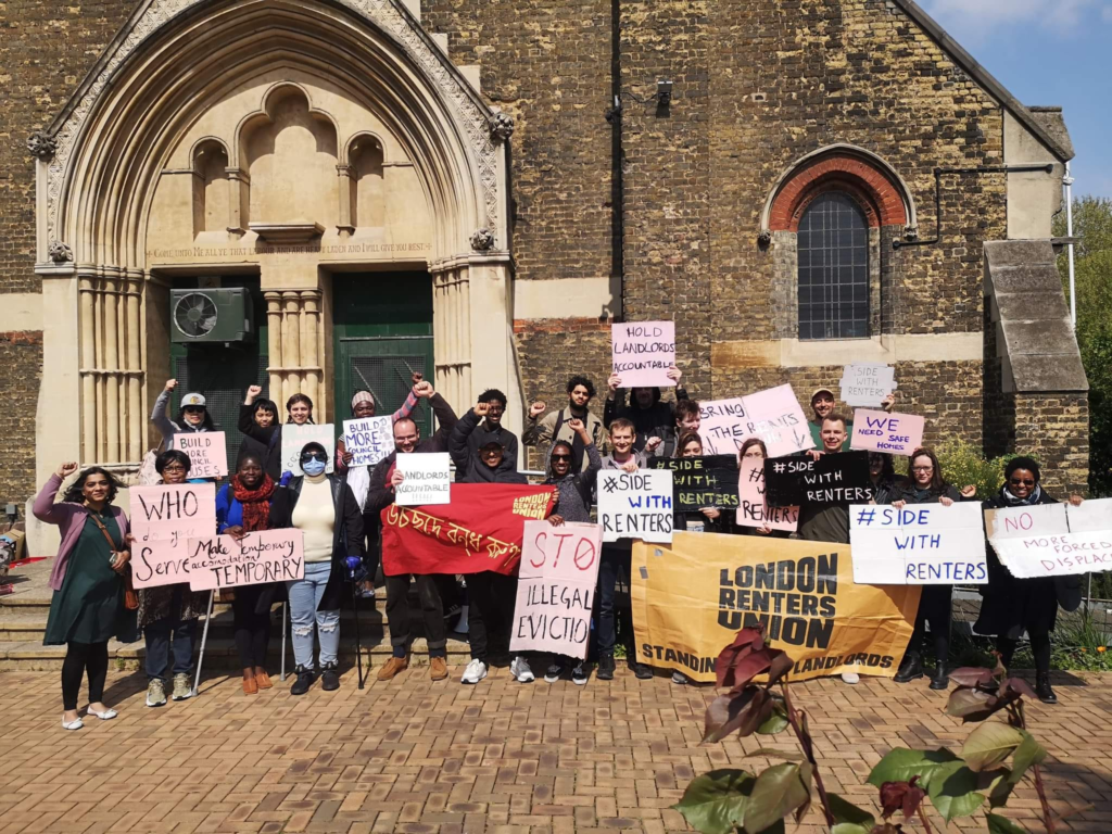 A multiracial group of renters in London holds pink and white signs with messages for renters' rights and stopping evictions. 
