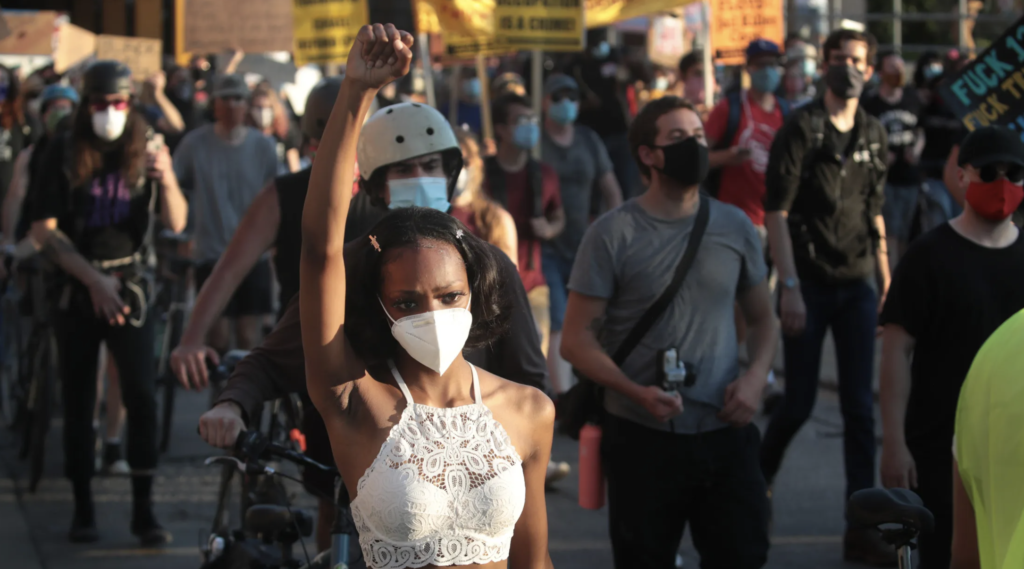 At a racial justice protest, a Black woman stands with her fist raised in a crowd of protesters. 