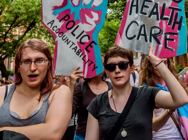Four trans gender activists hold up pink, white, and blue flags and signs for health care and police accountability during a march.