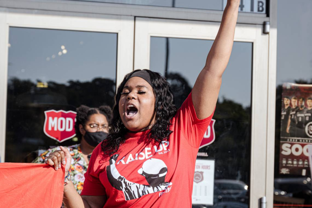 A Black woman in a red shirt holds a banner in one hand and calls out a message at a protest during a walkout against racism at work.