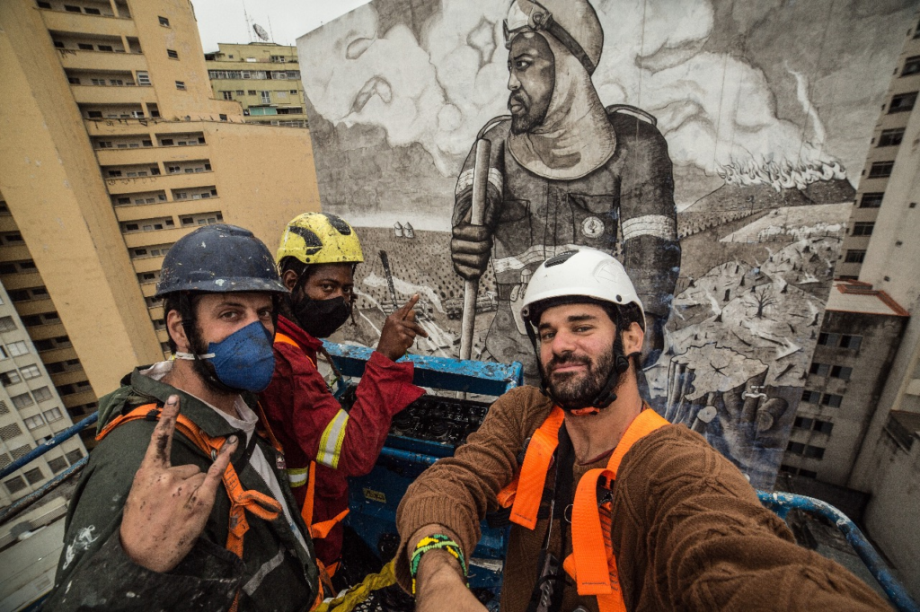 Three Brazilian artists working to protect the rainforest take a selfie in a construction lift in front of a skyscraper-sized mural made from the ashes of the rainforest.