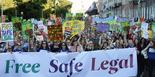 Irish women and allies with bright, colorful signs protest for reproductive rights. They hold a large banner that reads "free, safe, legal". 