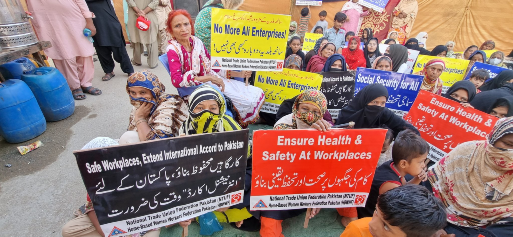 Pakistani women in headscarves sit with signs calling for workplace health and safety measures on 10th anniversary of factory fire.