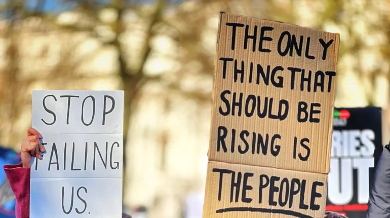 At a cost-of-living protest, a cardboard sign reads: the only thing that should be rising is the people.