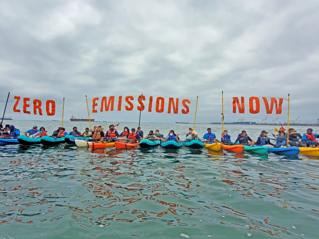 A line of activists in kayaks hold up banners that read: Zero Emissions Now.