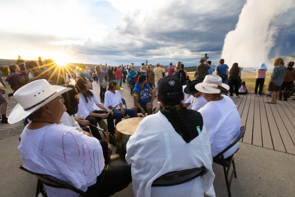 As part of an effort to re-indigenize the national park, Indigenous People drum in a circle while Yellowstone National Park's geyser erupts. 