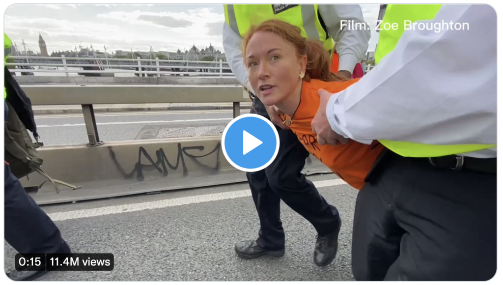 A handcuffed mother and activist in an orange tee-shirt gives an interview about the climate crisis while being hauled away by four police officers.