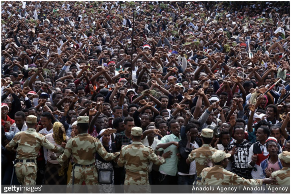 A large crowd Oromo people in Ethiopia lift crossed hands above their heads in a ritual that is also a protest against state repression. A group of police in uniform stand in front of the crowd.