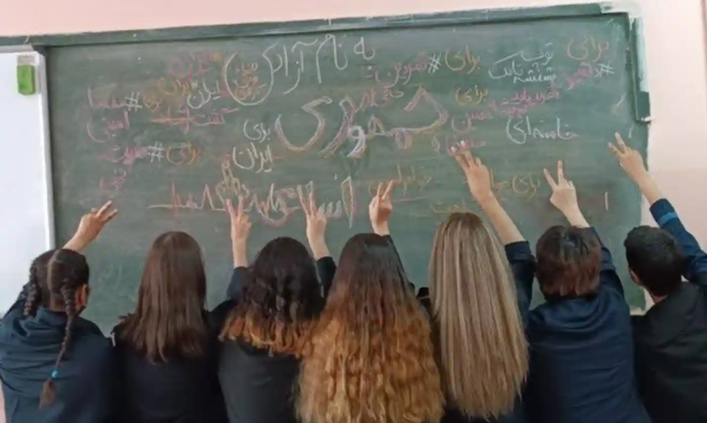 School girls in Iran raise peace signs in front of a chalkboard with writing on it. Their hair is uncovered as a protest against hijab laws and the recent murder of women by the Morality Police.