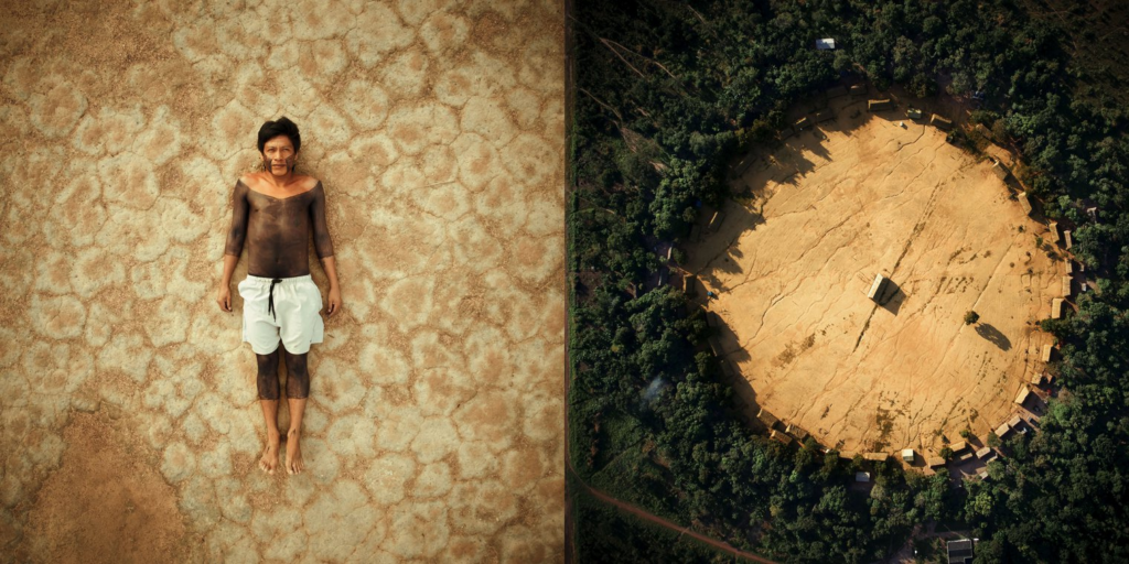 An Indigenous man in Brazil lies in the dirt in the center of a ring of houses in the rainforest. 