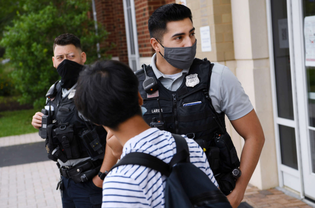 A student of color cranes to look at two school security officers who stand outside the front doors of his school. The officers were body armor and pandemic masks.