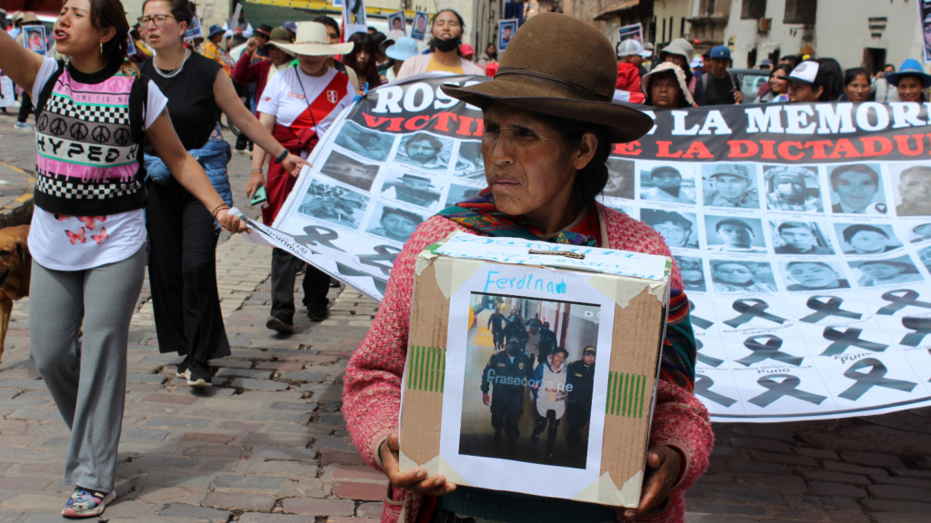 A Peruvian woman carries a box with a photo of a massacred protester. Behind her, a group bears a large banner with photos of others killed in a police crackdown on protests.