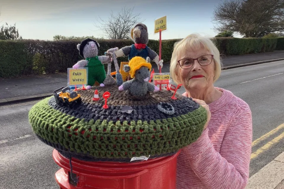 An elderly woman stands next to a hand-knit sculpture that sits atop a postbox and playfully protests the slow progress of the road construction.