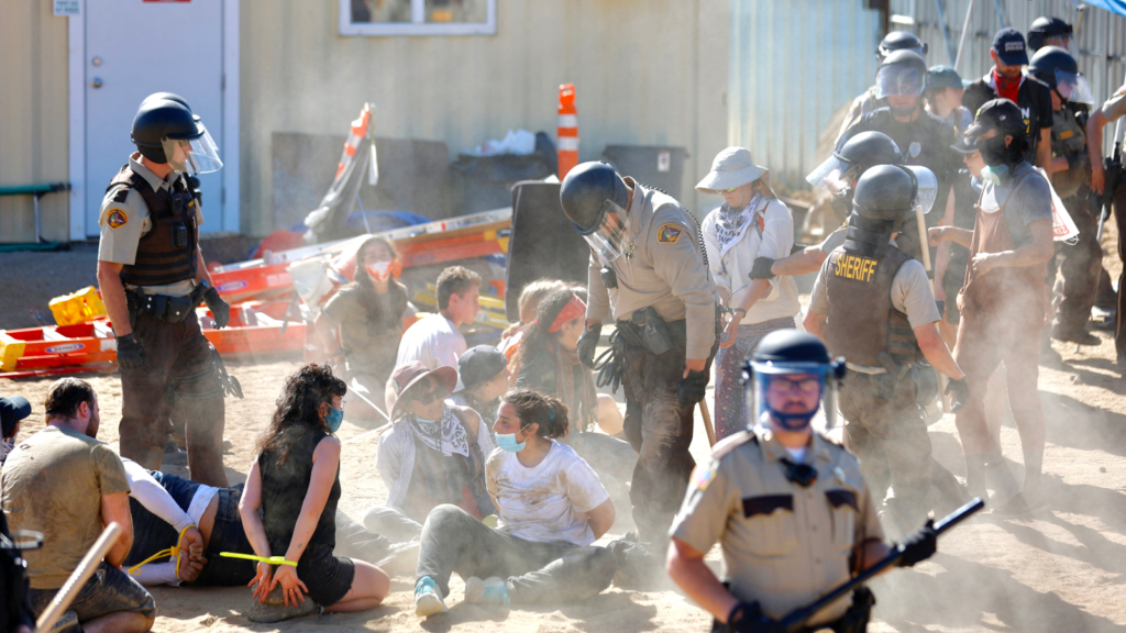 Police in helmets and body armor arrest climate activists who sit on the ground with hands zip-tied behind the back. 