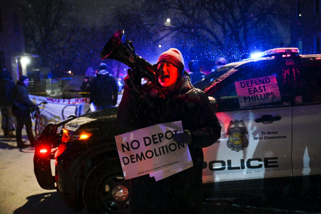 A protester speaks into a megaphone while standing in front of a police car in a snow storm. She holds a sign opposing the depot demolition.