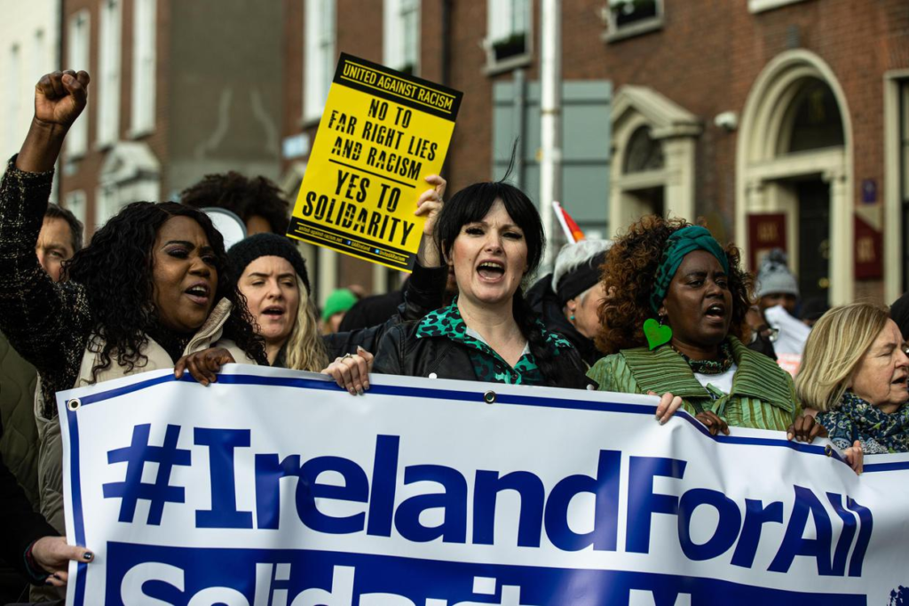 Black and other Irish citizens carry a large banner that reads #IrelandForAll as they show support for refugees.