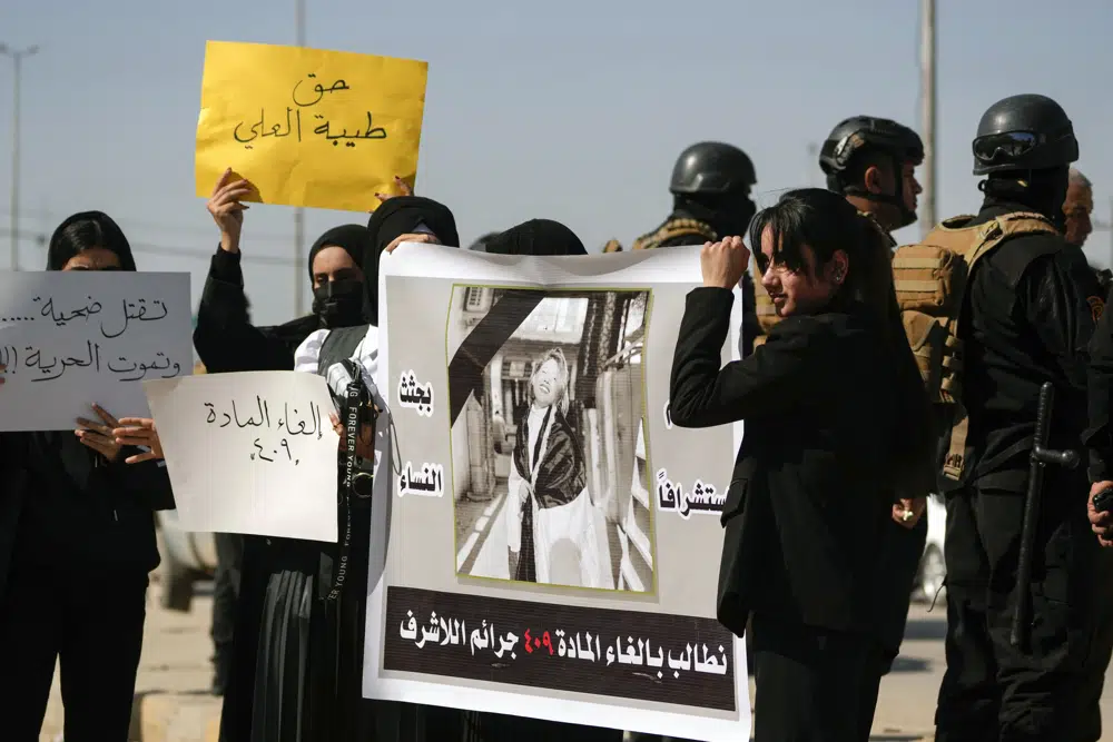 Iraqi women hold placards and a poster with a picture of Tiba Ali, a YouTube star who was recently killed by her father. A row of police stand behind them.