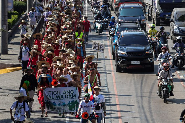 A group of Filipinos in red robes and wide-rimmed hats march on one side of the busy urban street to protest the construction of a dam.