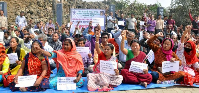A group of Indian women and men sit on a blue tarp on the side of the road chanting against an eviction campaign. They are members of a street vendors union.