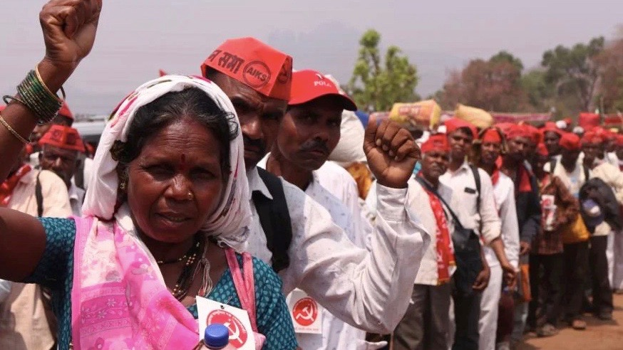 Wearing a sari, an Indian woman lifts her fist. Behind her a row of farmers where red hats as they protest.