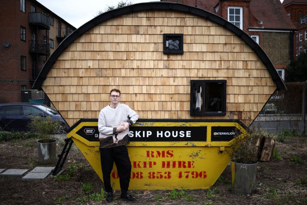 A man stands in front of the small house he built in a garbage dumpster in protest of high housing prices in London.