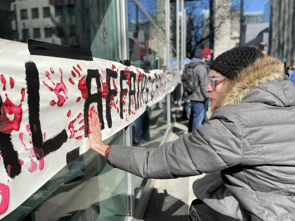 A long banner with red handprints is taped to the glass front of a Canadian bank. A peace protester adds her hand print to the image, demanding an end to arms trade in Yemen War.