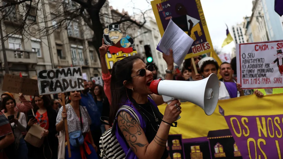 A woman holding leaflets and a large megaphone speaks to a crowd of protesters holding signs against high rents in Portugal.