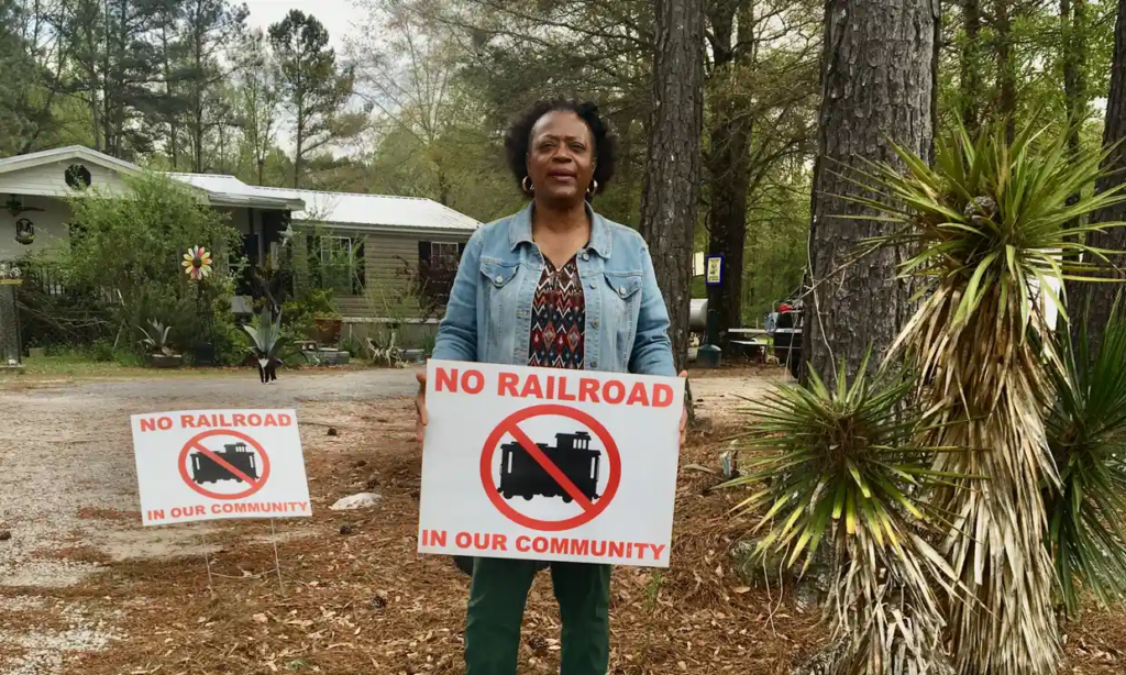 Near her home in the trees of Georgia, a Black activist holds a sign protesting a plan for a toxic railway line.