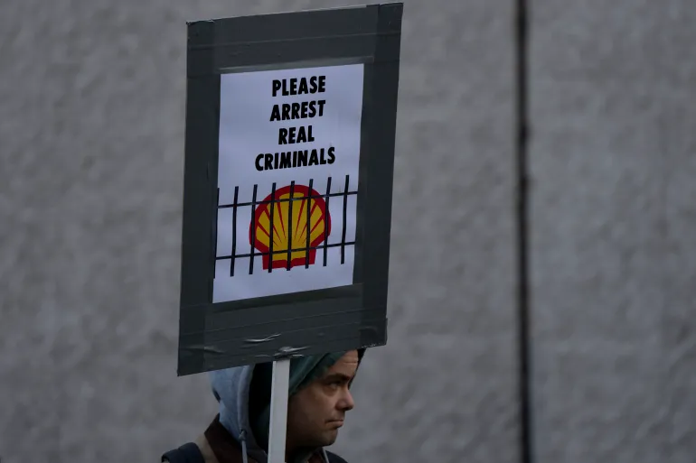 A Dutch climate protester holds a sign that reads: please arrest the real criminals. It shows a graphic of the Shell Oil Company logo behind bars.