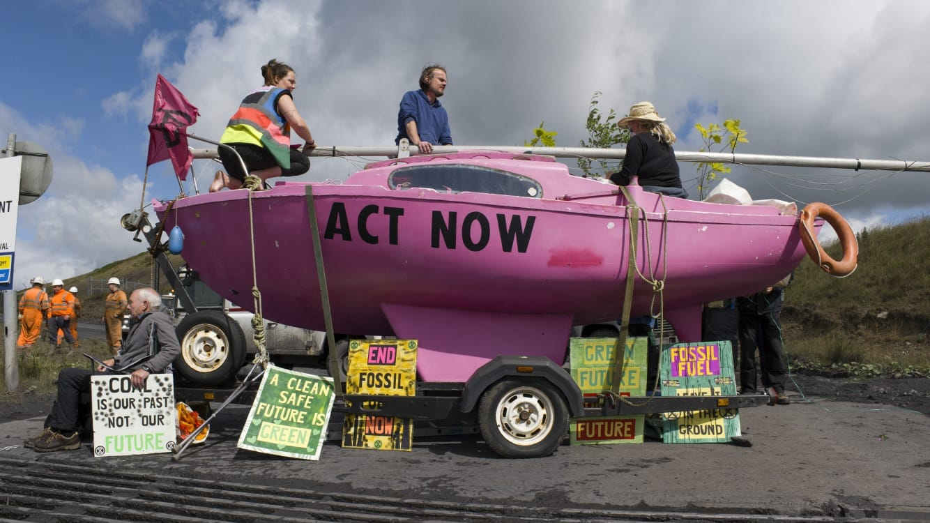 A bright pink sailboat with the words "act now" on the sides forms a barricade of a coal mine road in Wales. Activists sit on top and alongside it with colorful signs calling for an end to fossil fuel use. 