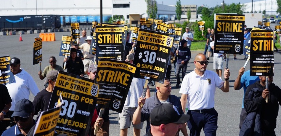 20+ Amazon workers in company shirts carry black and yellow signs on a picket line outside a warehouse.