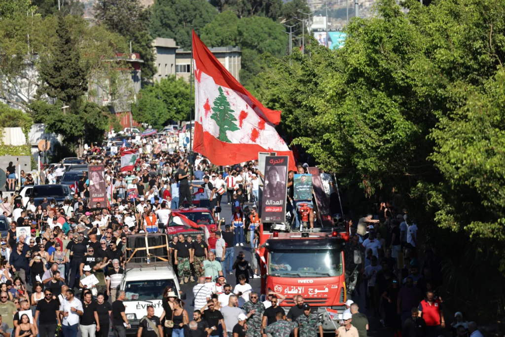 Hundreds march down a tree-lined street in Lebanon. Trucks carry memorial signs. A huge, red and white flag waves above the crowd.